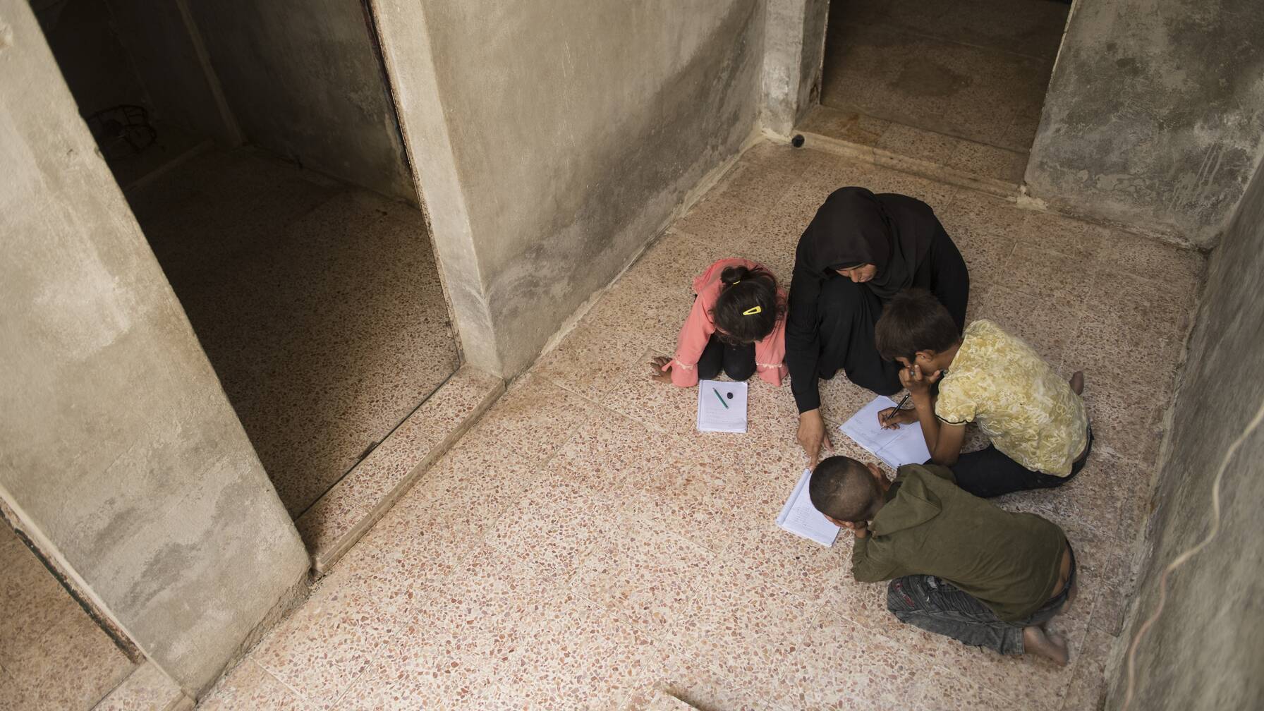 Fatima aide ses enfants à faire leurs devoirs. Elle n’a pas encore eu les moyens d’acheter une table et des chaises. La cage d'escalier est le meilleur éclairage - il n'y a pas d'électricité dans la maison.