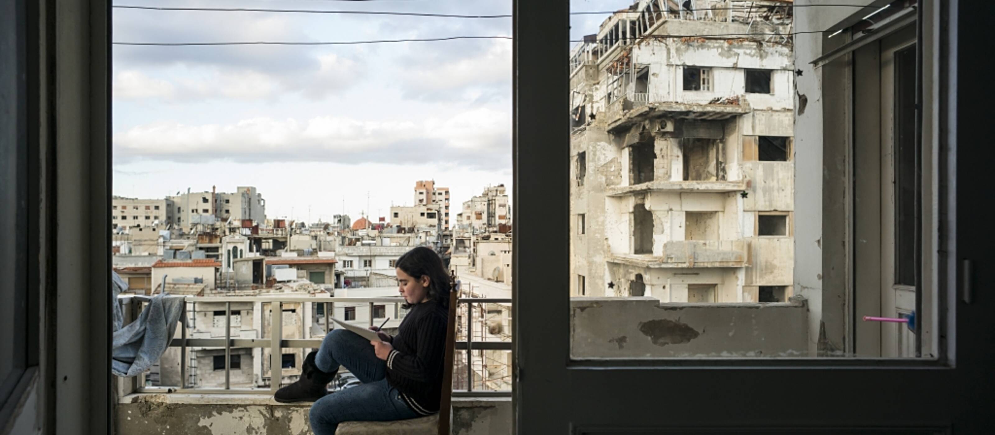 Ein Kind sitzt auf einem Stuhl auf dem Balkon und erledigt seine Hausaufgaben. Im Hintergrund ist die vom Krieg zerstörte Stadt zu sehen.