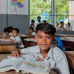 Ein Kind schaut inmitten seiner Klasse von seinen Schulbüchern auf und direkt in die Kamera.