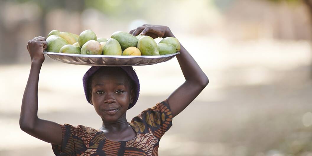 Eine Person trägt eine Schale voll frisch geernteter Mango und schaut direkt in die Kamera.