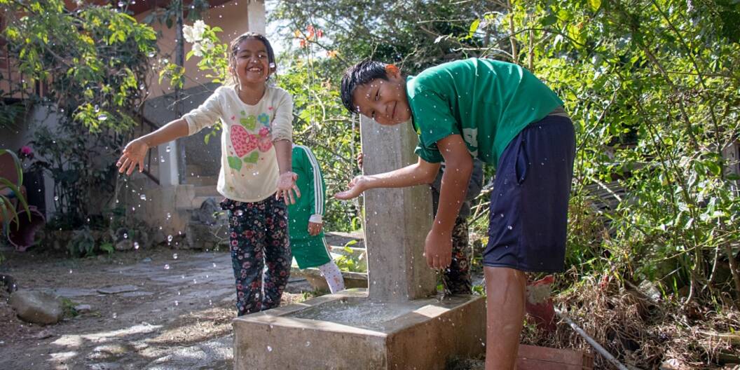 Zwei Kinder spritzen fröhlich Wassertropfen eines Brunnens in die Kamera.