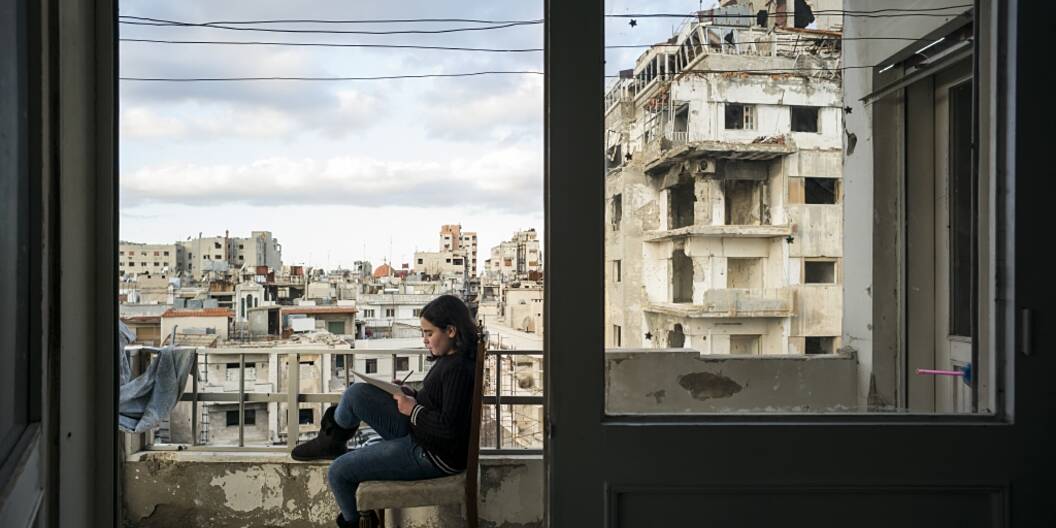 Ein Kind sitzt auf einem Stuhl auf dem Balkon und erledigt seine Hausaufgaben. Im Hintergrund ist die vom Krieg zerstörte Stadt zu sehen.